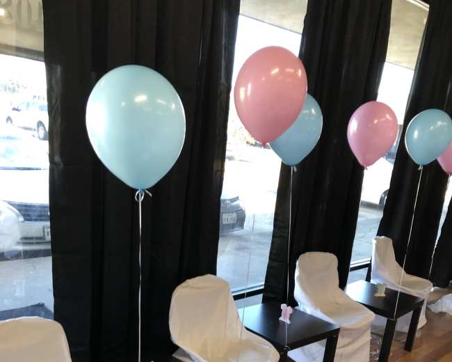 A row of four chairs covered in white cloth is set up next to a window, each adorned with pastel blue and pink balloons.