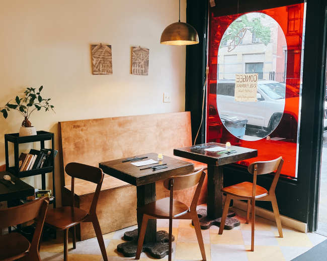 The image shows a small café interior with wooden tables and chairs, a large round red window, and minimalist decor.