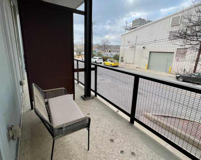 The image shows a narrow balcony with a metal-framed chair and a view of a street lined with parked cars and a building in the background.