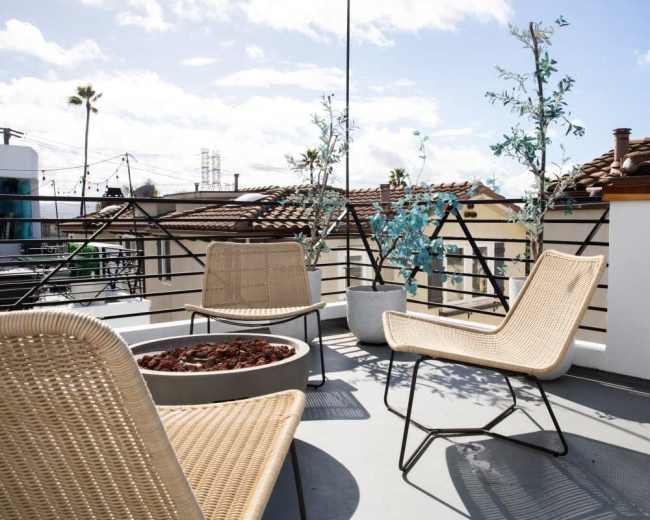 The image shows a rooftop terrace with three woven chairs, a round fire pit surrounded by small rocks, and two potted trees against a backdrop of a blue sky.