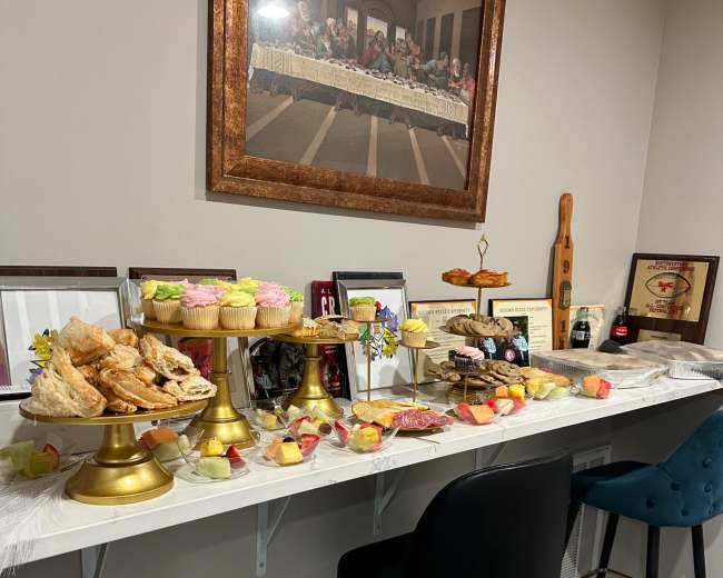 A long table displays an assortment of desserts, including pastries, cupcakes, and fruit, arranged with decorative items and framed pictures.