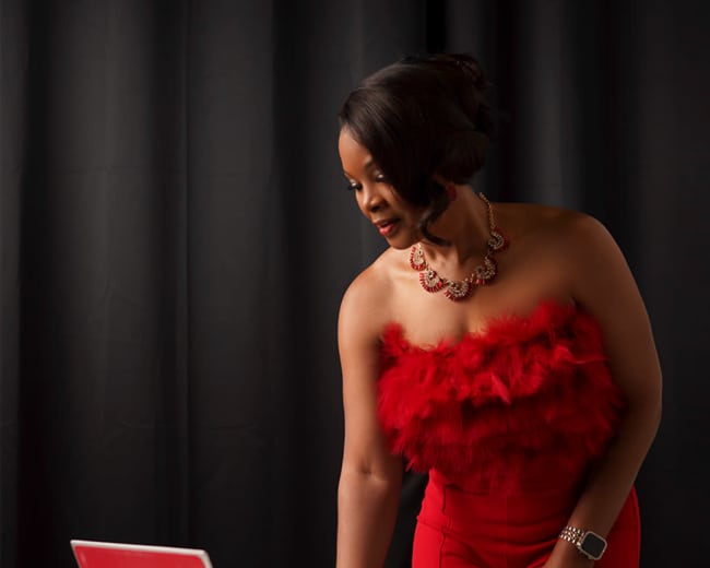 A woman in a red outfit stands beside a laptop on a white table, examining the screen.