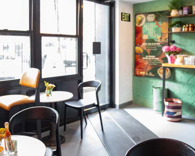 The image shows a cozy café interior with small tables, black chairs, and a decorative green wall featuring promotional artwork.