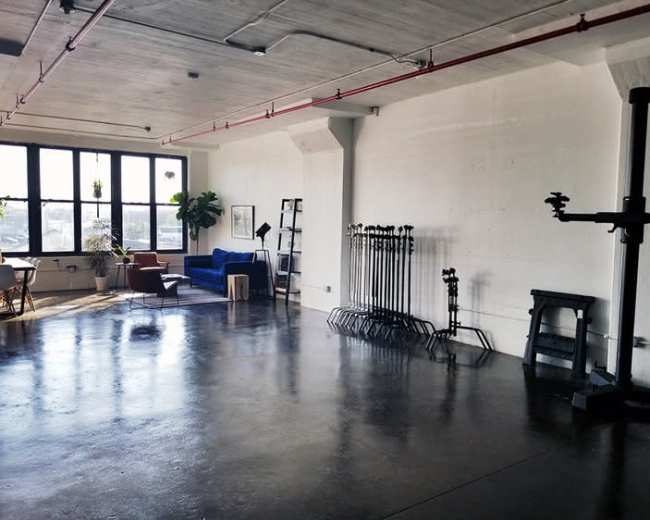 An industrial-style interior space with large windows, a blue couch, several plants, and multiple photography stand supports along the wall.