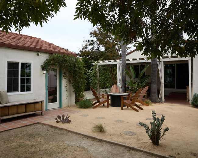 The image shows a cozy outdoor space featuring a patio with wooden chairs, a small table, and surrounding greenery near a house with a red-tiled roof.