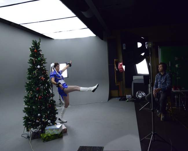A person in a blue outfit poses playfully in front of a decorated Christmas tree while another person captures the moment with a camera.