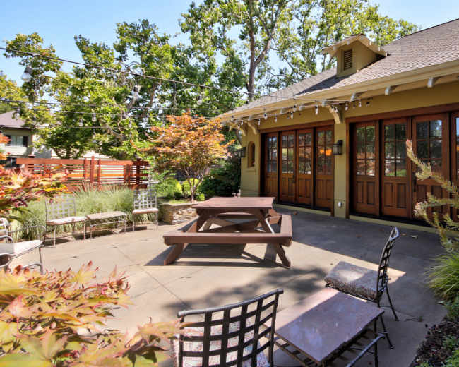 The image shows a patio area with a wooden picnic table, several chairs, and landscaped greenery, adjacent to a house with large glass doors.