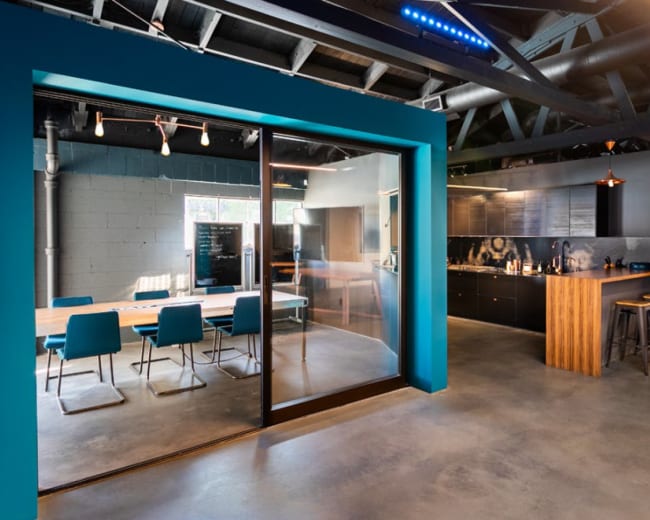 A modern office space features a dining area with a long table and blue chairs, adjacent to a kitchen with dark cabinetry and wooden accents.