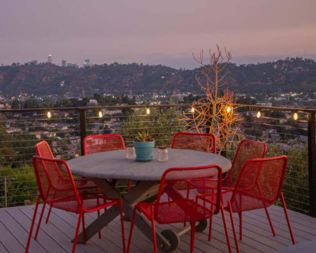 A round table surrounded by red chairs is set on a wooden deck with city lights in the distance against a twilight sky.