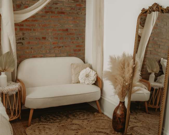 The image shows a cozy corner of a room featuring a light-colored loveseat, a tall mirror with an ornate frame, and decorative elements like a vase of dried flowers and a textured rug against a brick wall.