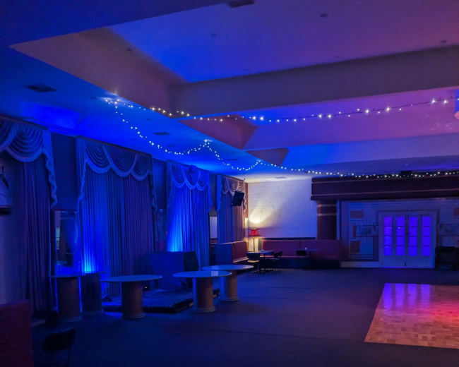 The image depicts a dimly lit room with blue lighting, featuring curtains, a wooden floor, and scattered furniture.