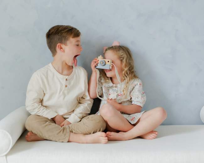 A boy sits next to a girl on a white sofa, with the girl playfully taking a photo using a vintage camera while the boy sticks out his tongue.
