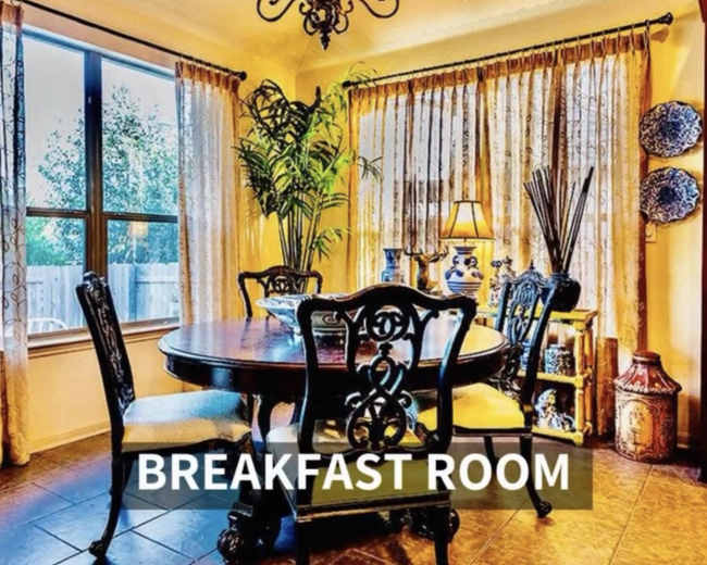 A breakfast room features a round table surrounded by chairs, illuminated by a chandelier, with large windows and decorative plants.