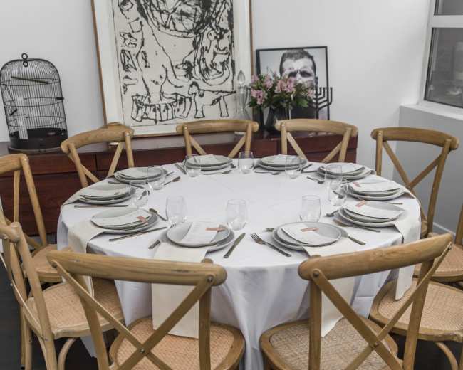A round dining table is set with plates and glasses, surrounded by wooden chairs, against a backdrop of artwork and a birdcage.