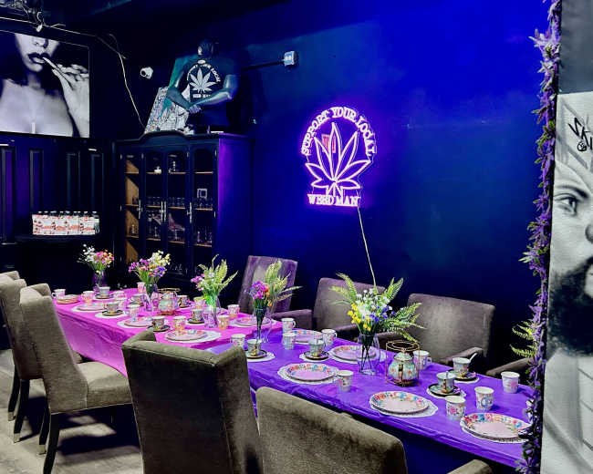 A vibrant dining setup is displayed, featuring a long table adorned with purple linens, floral centerpieces, and a neon cannabis-themed sign on a dark wall.