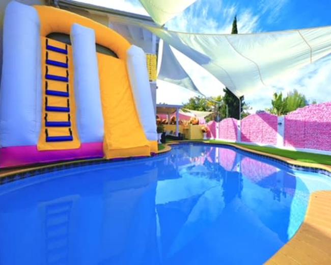 A colorful inflatable slide is positioned beside a clear blue pool, surrounded by manicured grass and a decorative pink wall.