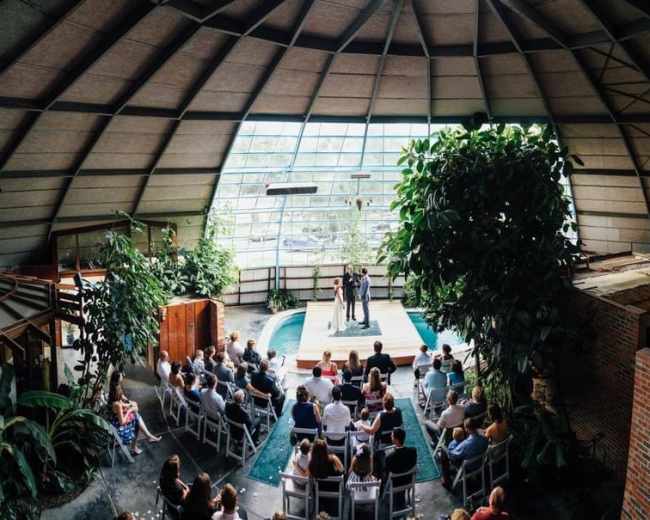 A wedding ceremony takes place indoors under a large, arched glass structure surrounded by tropical plants and a small pool.