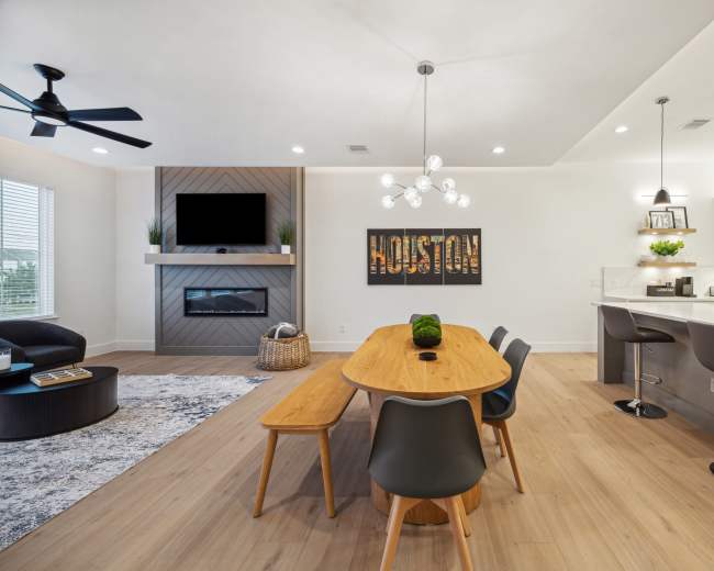The image shows a modern living space with a dining table and chairs, a sectional couch, and a wall-mounted TV above a fireplace.