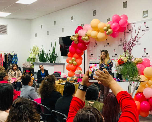 A speaker stands in front of a decorated backdrop with balloons and floral arrangements, addressing an audience seated in a brightly lit room.