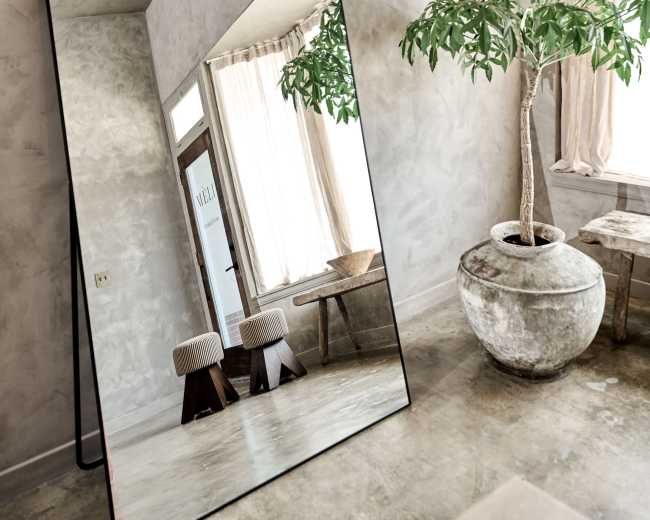 A large, arched mirror reflects a room with a potted plant, wooden stools, and natural light filtering through sheer curtains.