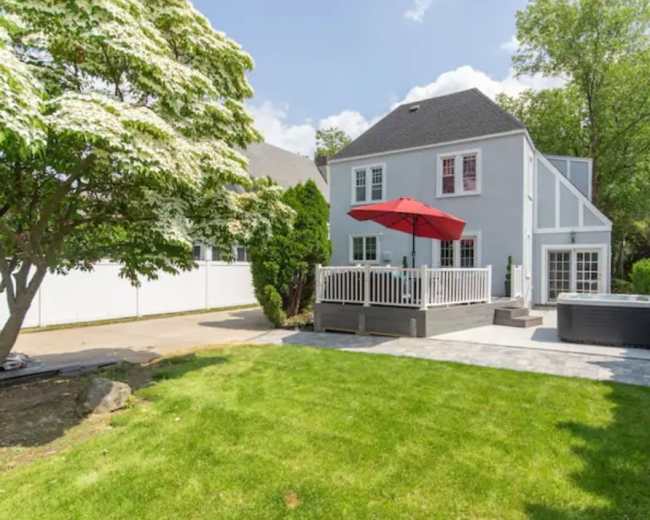 The image shows a house with a gray exterior, a deck featuring a red umbrella, and a hot tub, all set in a well-maintained yard with green grass and trees.