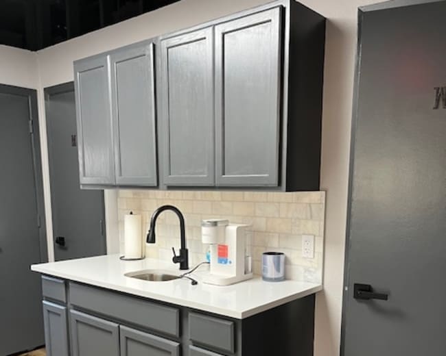 The image shows a small kitchen area with gray cabinets, a white countertop, a sink, and a few appliances.