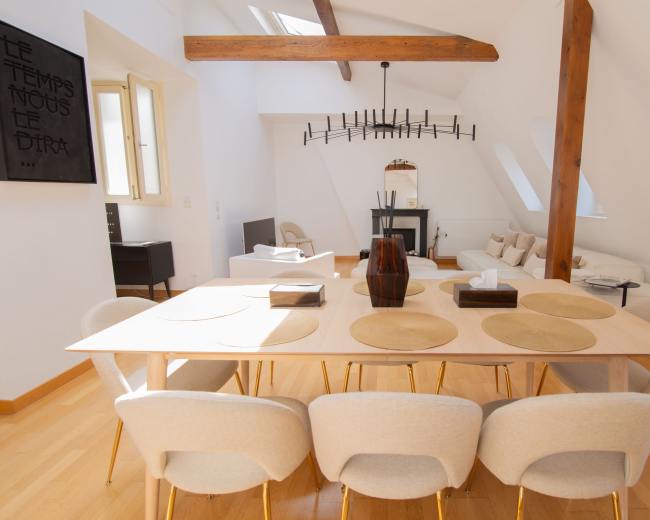 A spacious, well-lit living area features a wooden dining table surrounded by upholstered chairs, with a modern fireplace and seating area in the background.