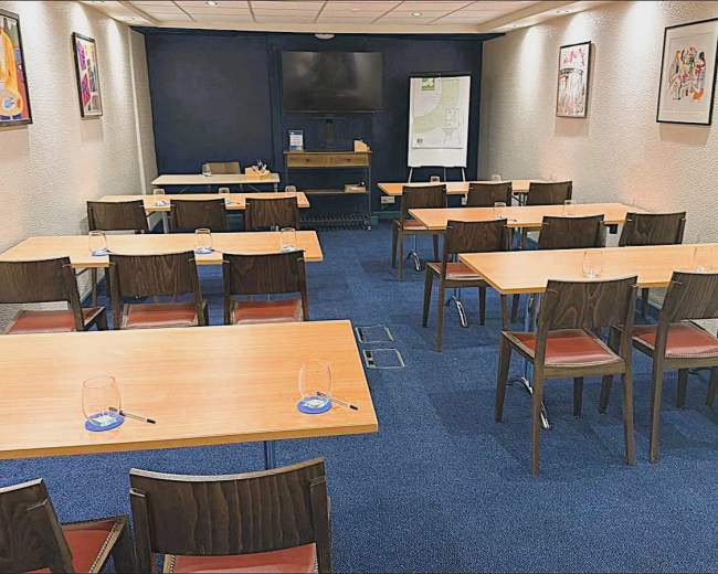 The image shows a conference room with wooden tables and chairs arranged in a grid format, with a television on the wall and artwork displayed.