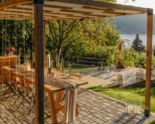 A wooden pergola frames a dining setup with tables and chairs, overlooking a scenic landscape featuring greenery and a body of water in the distance.