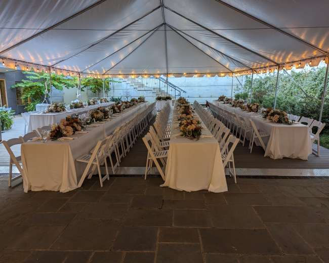 A large outdoor tent is set with multiple tables covered in white tablecloths, arranged in two parallel lines, each adorned with flower centerpieces.