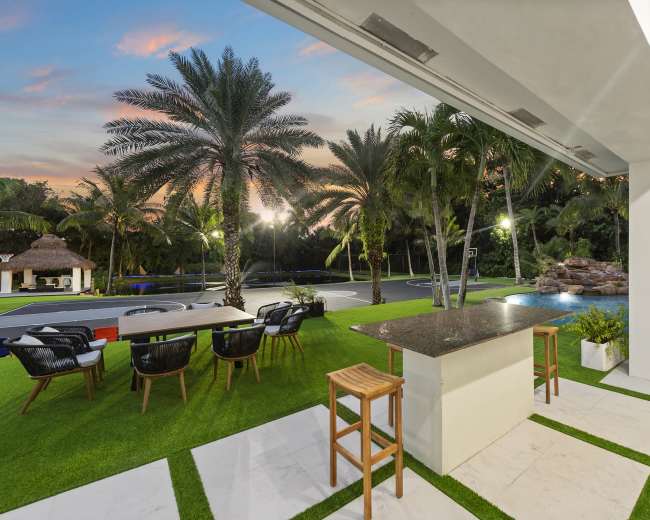 The image shows an outdoor kitchen area with bar seating, surrounded by palm trees and a basketball court, against a backdrop of a colorful sunset.