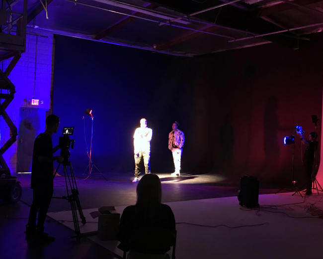 Two individuals are illuminated in a dimly lit studio, surrounded by various stage lights and camera equipment.