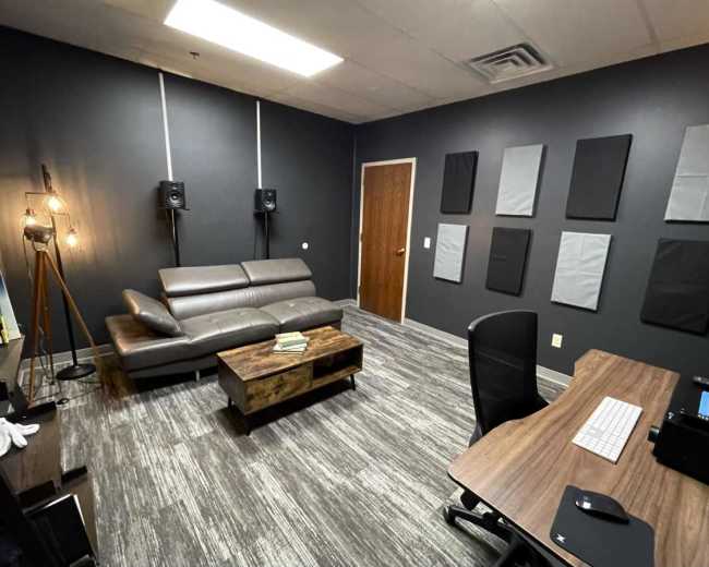 The image shows a modern office or studio space featuring a gray leather sofa, a wooden coffee table, soundproofing panels on the walls, and a desk with a computer.