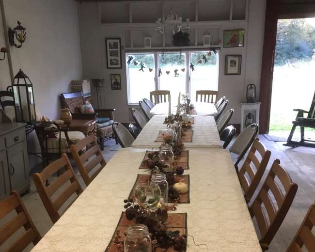 A long, rectangular dining table is set with lace runners and jars, arranged for a gathering in a spacious room with large windows.