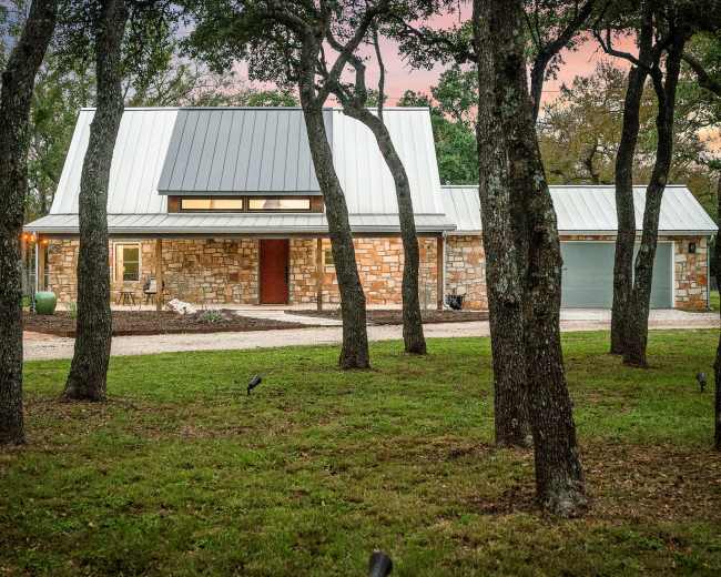 A modern stone house with a metal roof and a driveway is set among several trees at dusk.