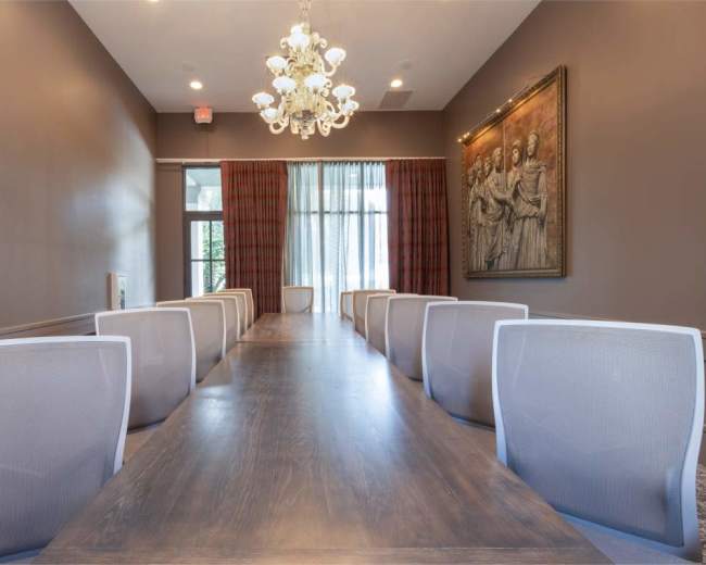 A long wooden conference table is set under a chandelier in a well-lit meeting room, with seating on either side and large windows at one end.