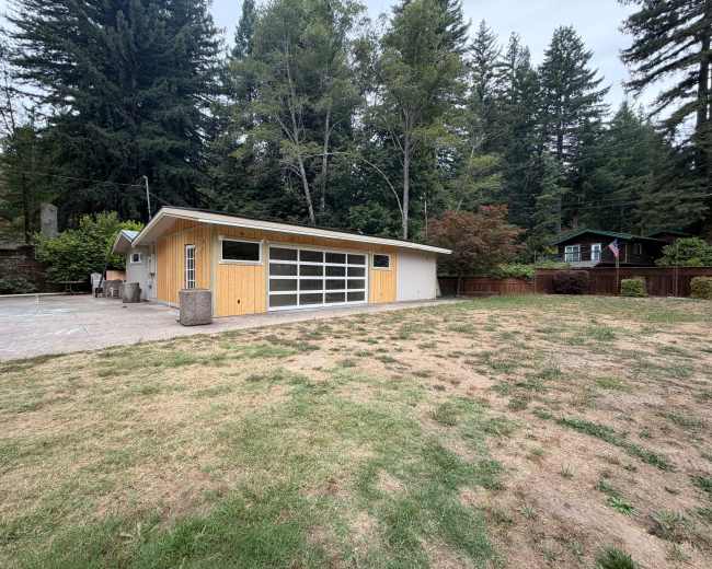 A modern single-story house with a large glass garage door sits on a grassy plot surrounded by tall trees and other homes in the background.