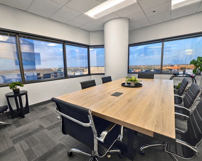 A modern conference room features a large wooden table surrounded by black leather chairs and floor-to-ceiling windows offering a view of the outside.