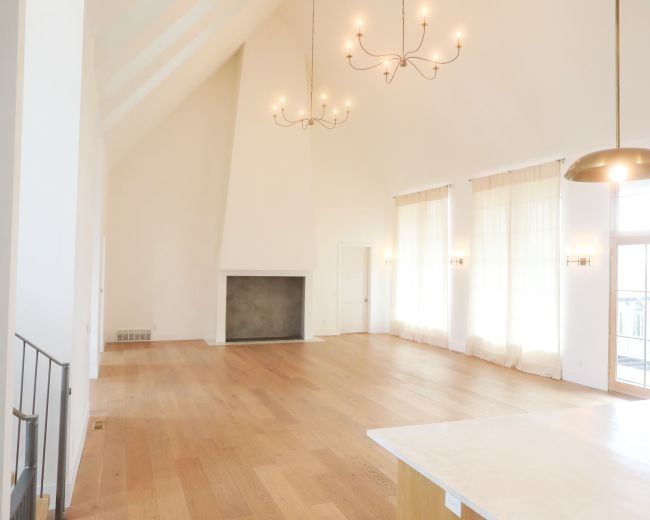 A large, bright living room features high ceilings, wooden flooring, and large windows with sheer curtains.