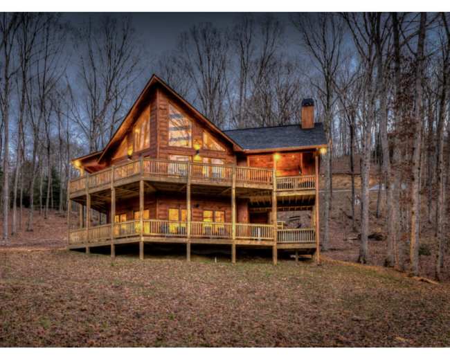 The image shows a large wooden cabin with multiple balconies set among bare trees in a wooded area during dusk.