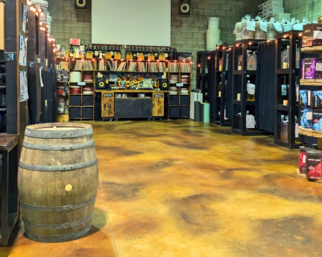 The image shows a spacious interior of a warehouse with shelves filled with various containers and a wooden barrel placed in the foreground.