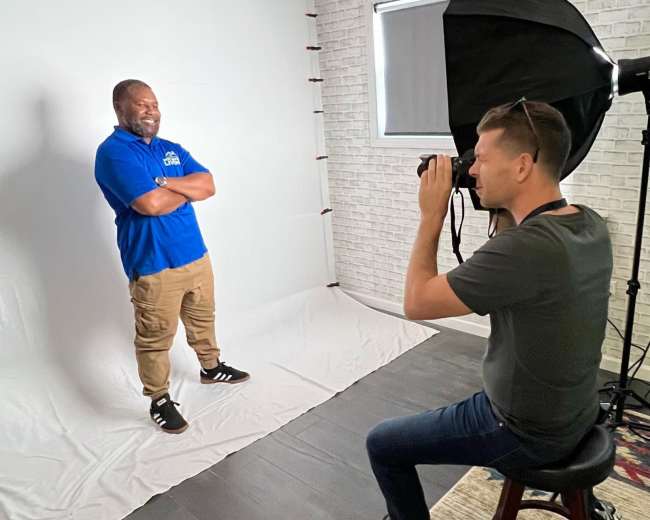 A photographer captures a smiling man in a blue shirt while he poses with his arms crossed in a studio setting with a white backdrop.