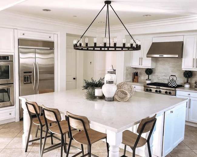 A modern kitchen features a large central island with wooden barstools, stainless steel appliances, and a circular chandelier above.