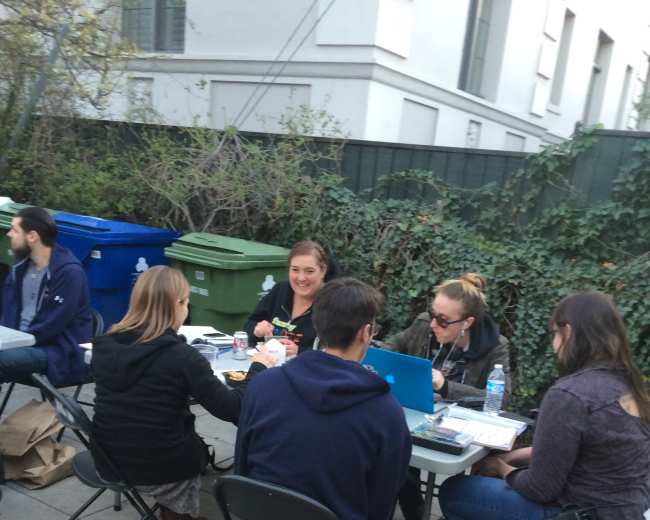 A group of six people is seated around a table outdoors, engaged in work or discussion with laptops and papers spread out.
