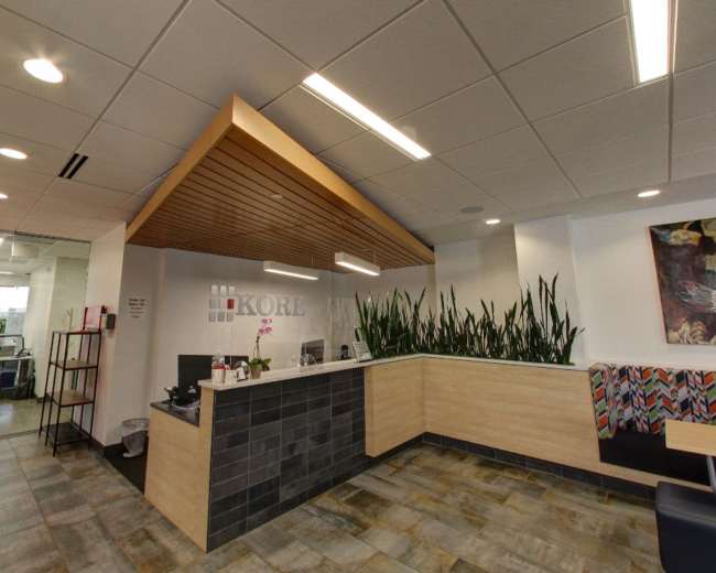 The image shows a modern reception area with a wooden ceiling feature, a sleek front desk, and green plants along the wall.