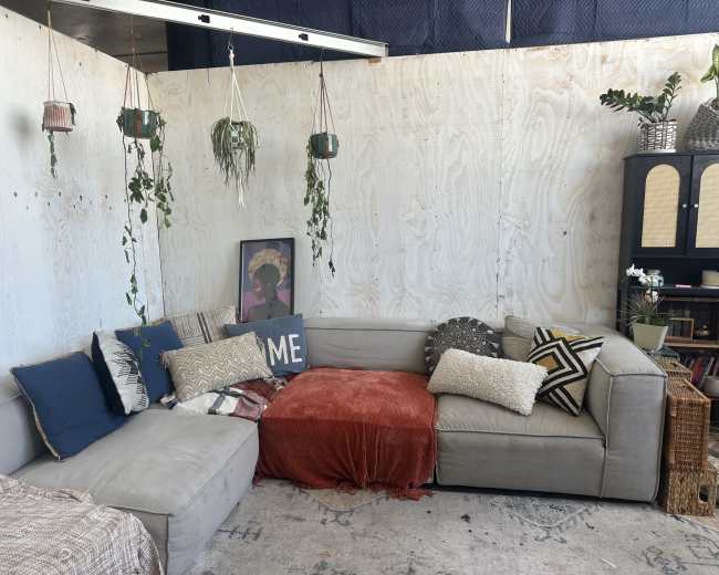 The image shows a cozy living area with a gray sectional sofa, various decorative pillows, a wooden plant stand, hanging plants, and a small bookshelf against a plywood wall.