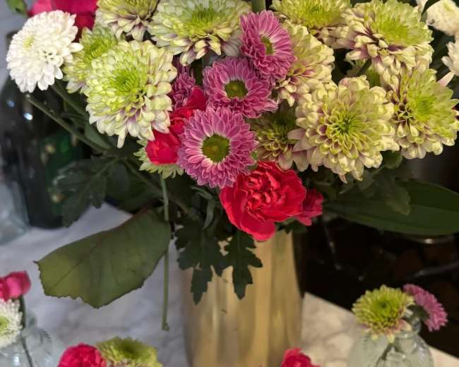 A large floral arrangement with pink, white, and green flowers is displayed in a gold vase, accompanied by small glass bottles containing individual blooms.
