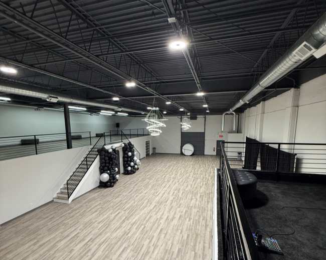 The image shows a spacious vacant interior with wood-like flooring, high ceilings, and a staircase leading to an upper level.