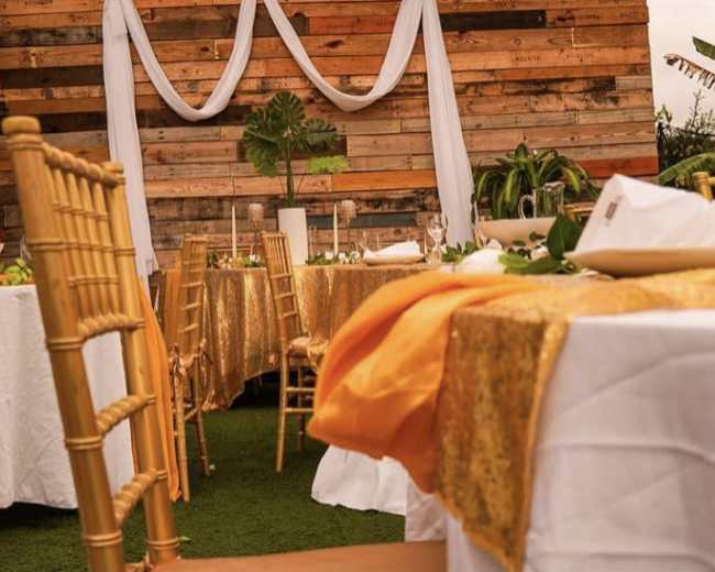 A decorated dining area features tables with white and gold tablecloths, surrounded by gold chiavari chairs, against a wooden wall.