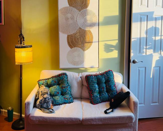 A white couch with patterned pillows is positioned against a green wall, illuminated by sunlight, with a decorative wall hanging and a door in the background.